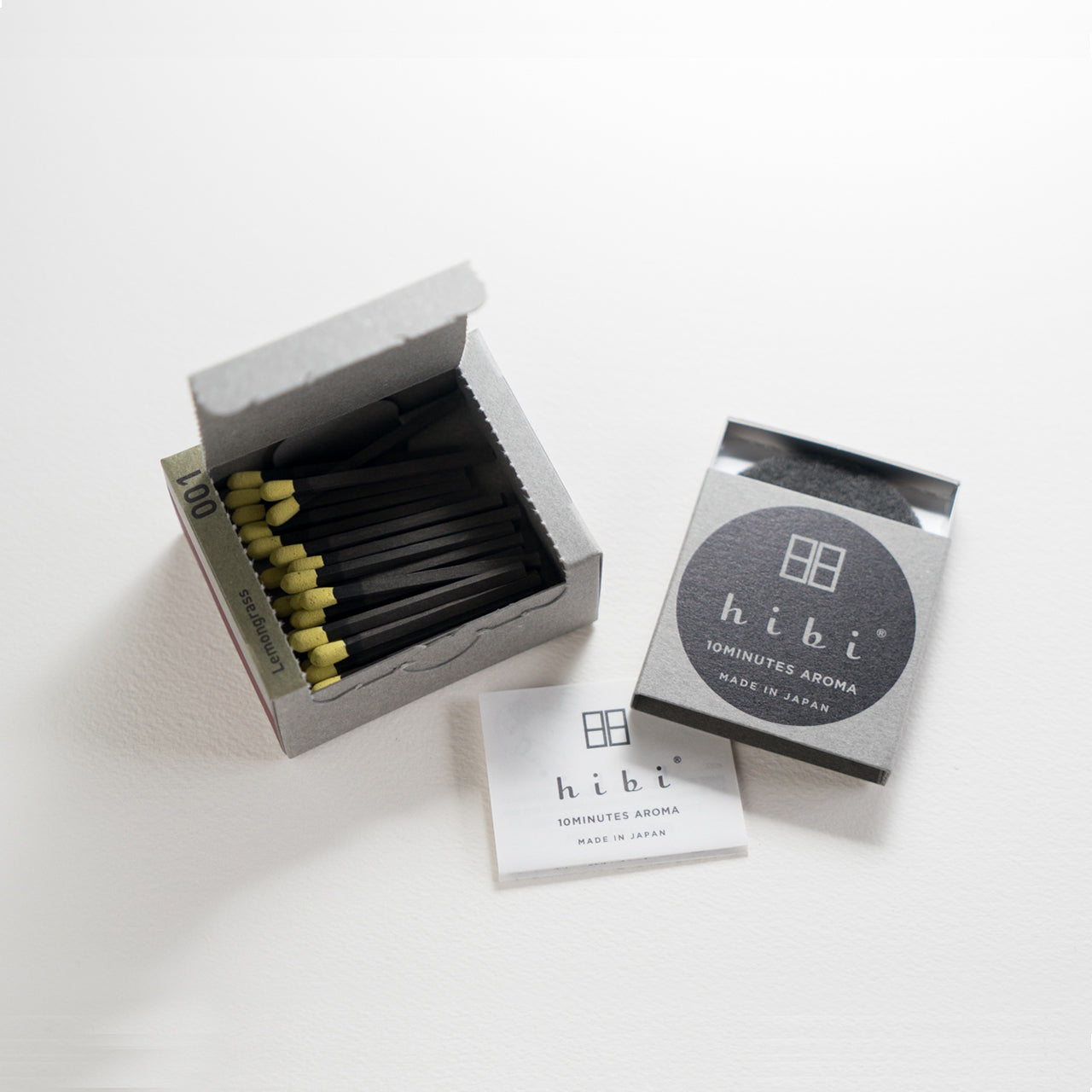 An opened box of Hibi 10 Minutes Aroma Lemongrass incense matches containing 30 incense sticks with yellow tips. Next to it is a box containing a non-flammable incense mat. All set in a white background.