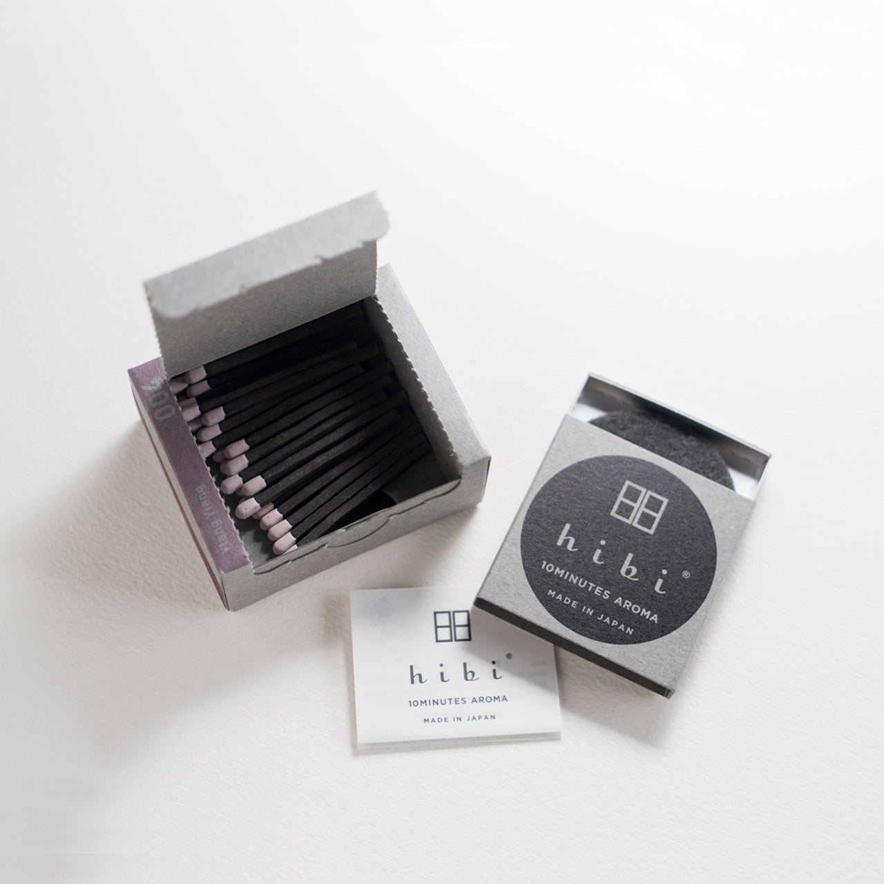 An opened box of Hibi 10 Minutes Aroma Ylang Ylang incense matches containing 30 incense sticks with yellow tips. Next to it is a box containing a non-flammable incense mat. All set in a white background.