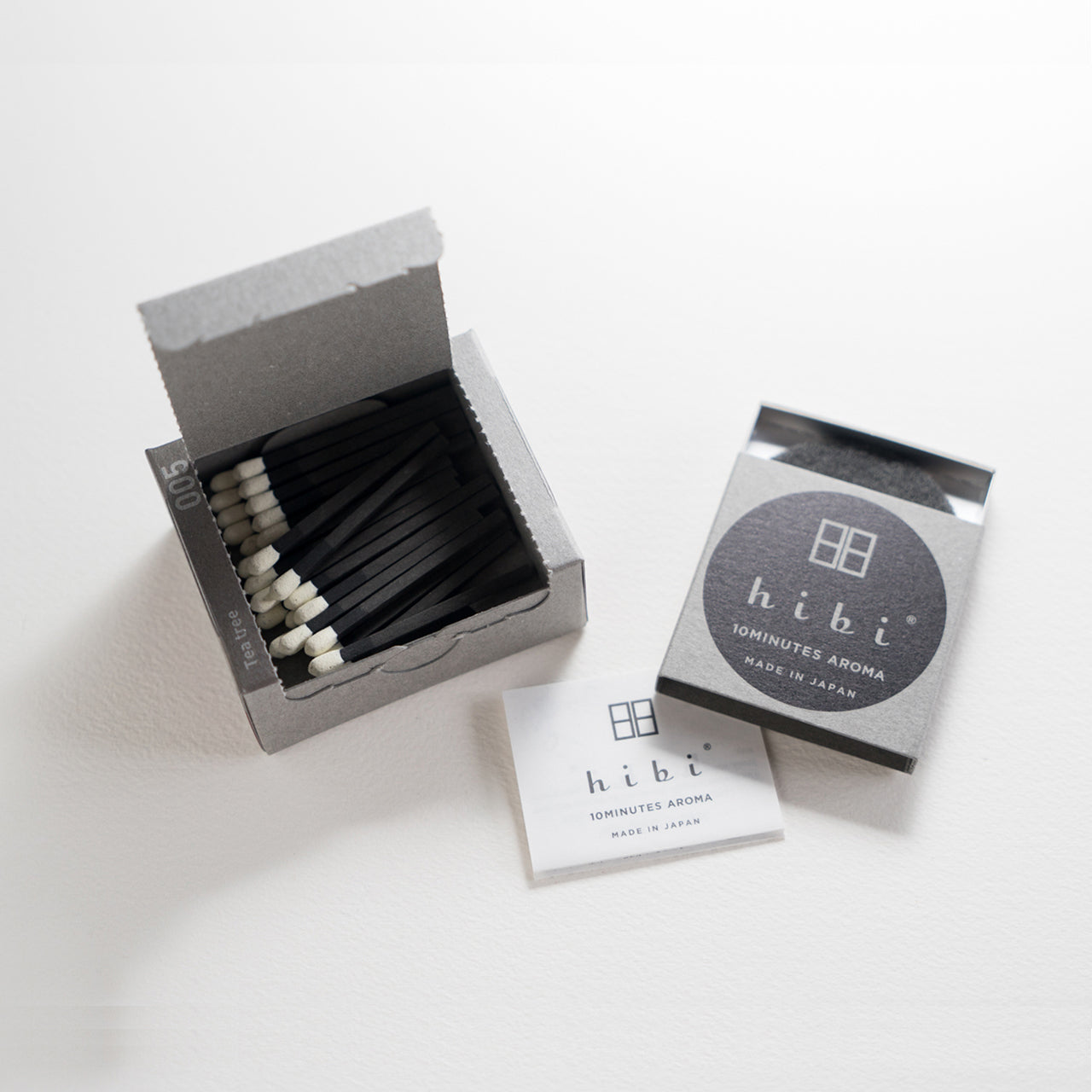 An opened box of Hibi 10 Minutes Aroma Tea Tree incense matches containing 30 incense sticks with yellow tips. Next to it is a box containing a non-flammable incense mat. All set in a white background.