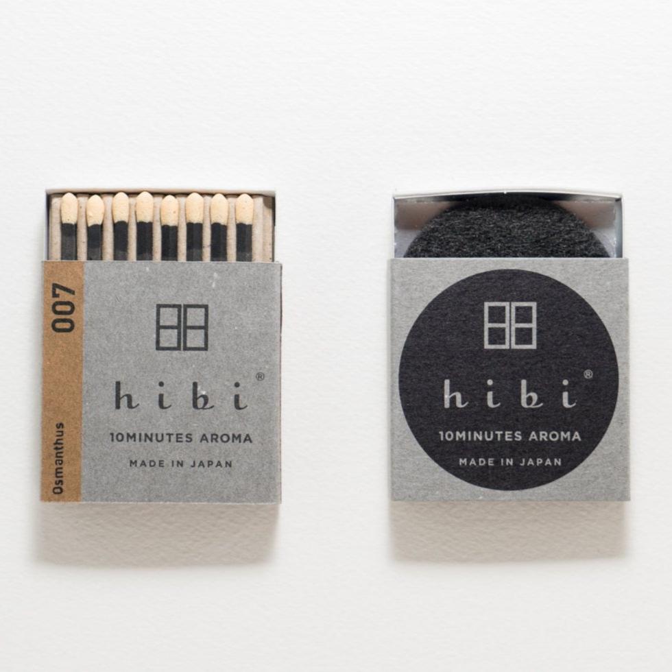 An opened box of Hibi 10 Minutes Aroma Osmanthus incense matches containing 8 incense sticks with yellow tips. Next to it is a box containing a non-flammable incense mat. All set in a white background.
