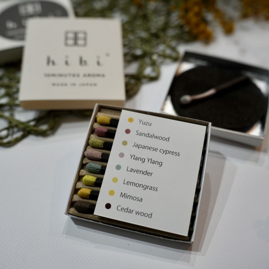 An opened box of Hibi 10 Minutes Aroma Special Edition incense matches containing 8 incense sticks with different colour tips. There is a sheet identifying the 8 incense sticks - Yuzu, Sandalwood, Japanese Cypress, Ylang Ylang, Lavendar, Lemongrass, Mimosa, Cedarwood. There are a burning incense stick laying on top of a non-flammable incense mat and a hibi incense box cover.
