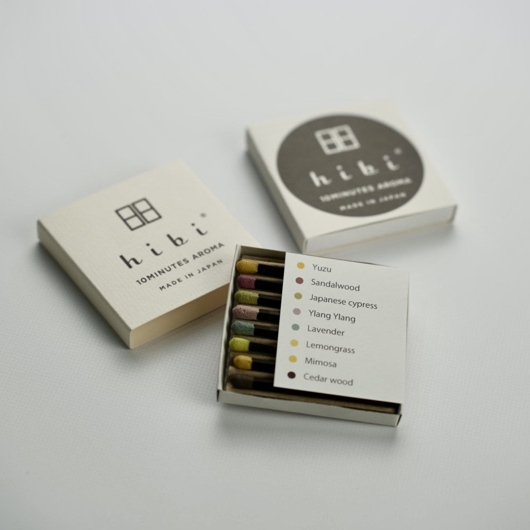 An opened box of Hibi 10 Minutes Aroma Special Edition incense matches containing 8 incense sticks with different colour tips. There is a sheet identifying the 8 incense sticks - Yuzu, Sandalwood, Japanese Cypress, Ylang Ylang, Lavendar, Lemongrass, Mimosa, Cedarwood. In the background are two boxes labeled as hibi 10 Minutes Aroma, Made in Japan.