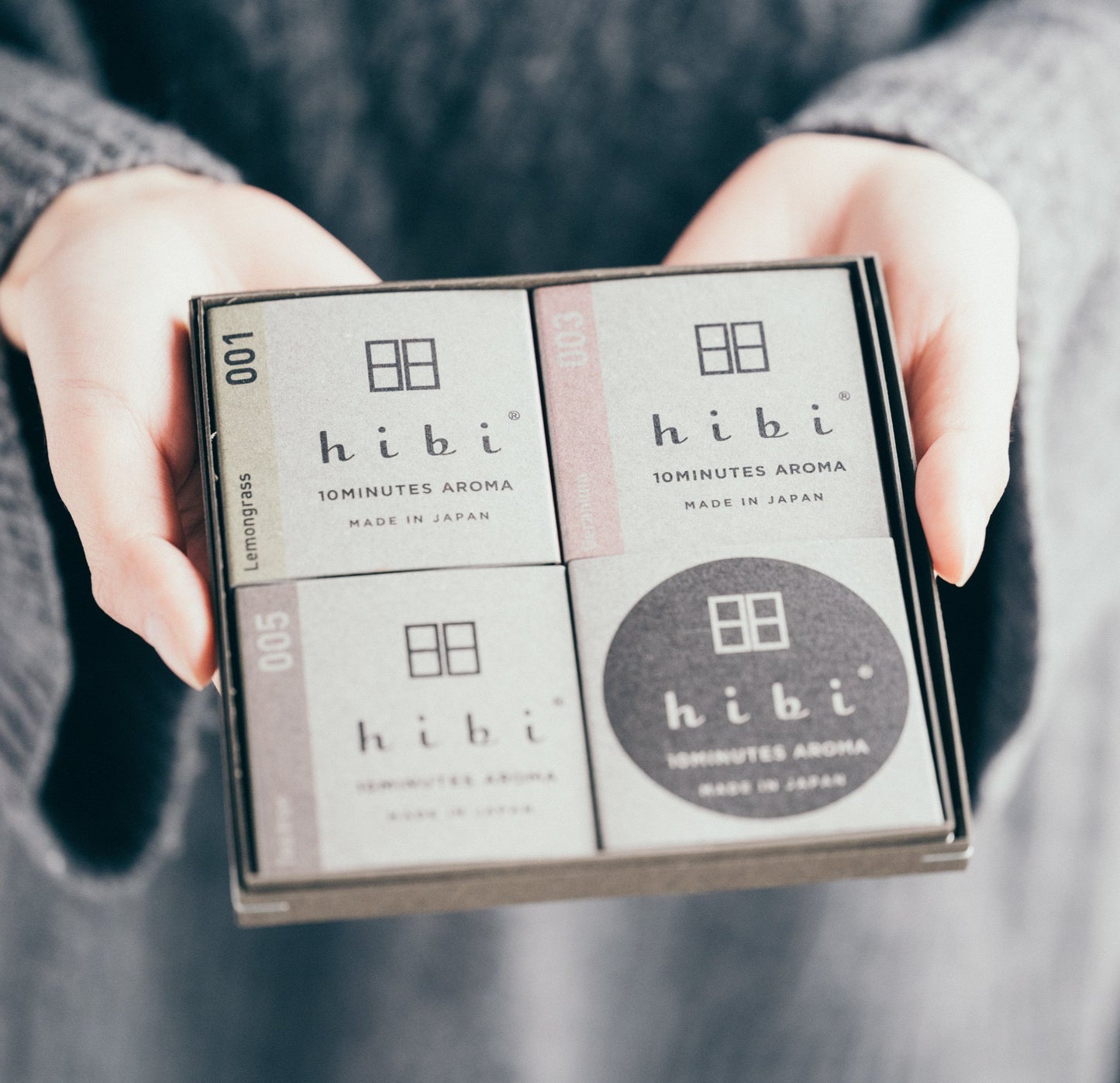 Two hands are holding an opened grey colour gift box which contains three packs of Lemongrass, Geranium, and Tea Tree incense matches, and a non-flammable incense mat.