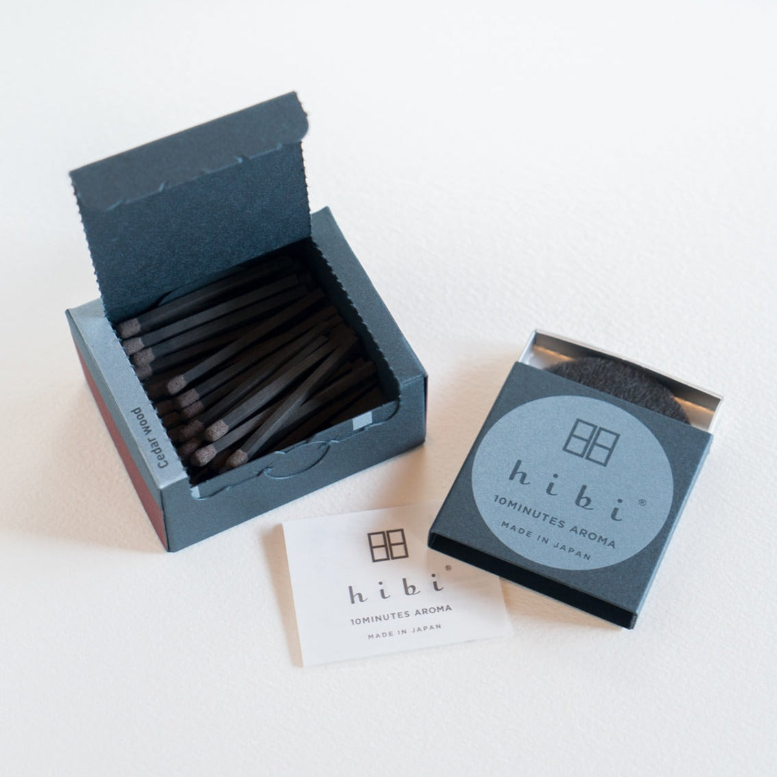 An opened box of Hibi 10 Minutes Aroma Cedar Wood incense matches containing 30 incense sticks with pink tips. Next to it is a box containing a non-flammable incense mat. All set in a white background.