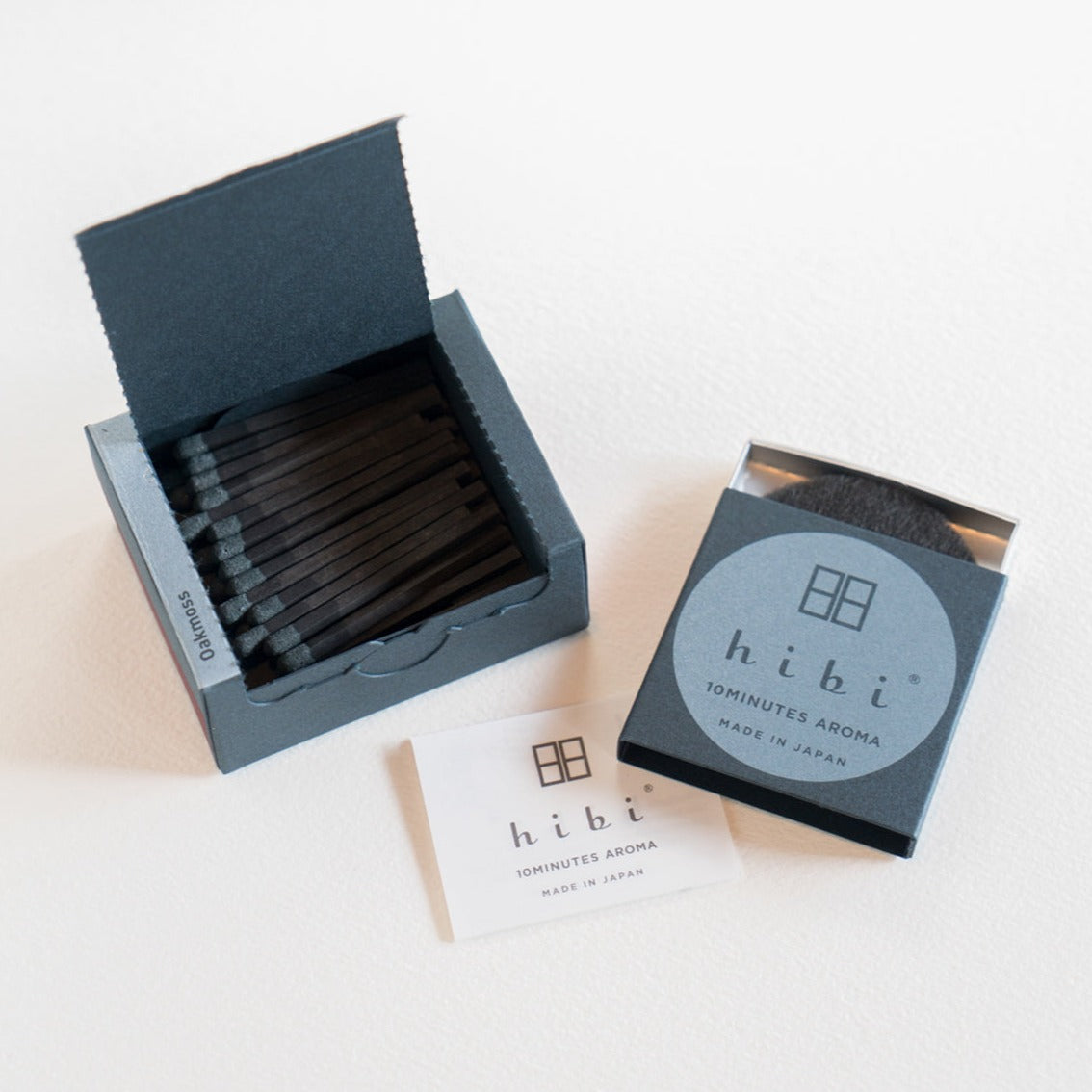 An opened box of Hibi 10 Minutes Aroma Oak Moss incense matches containing 30 incense sticks with pink tips. Next to it is a box containing a non-flammable incense mat. All set in a white background.