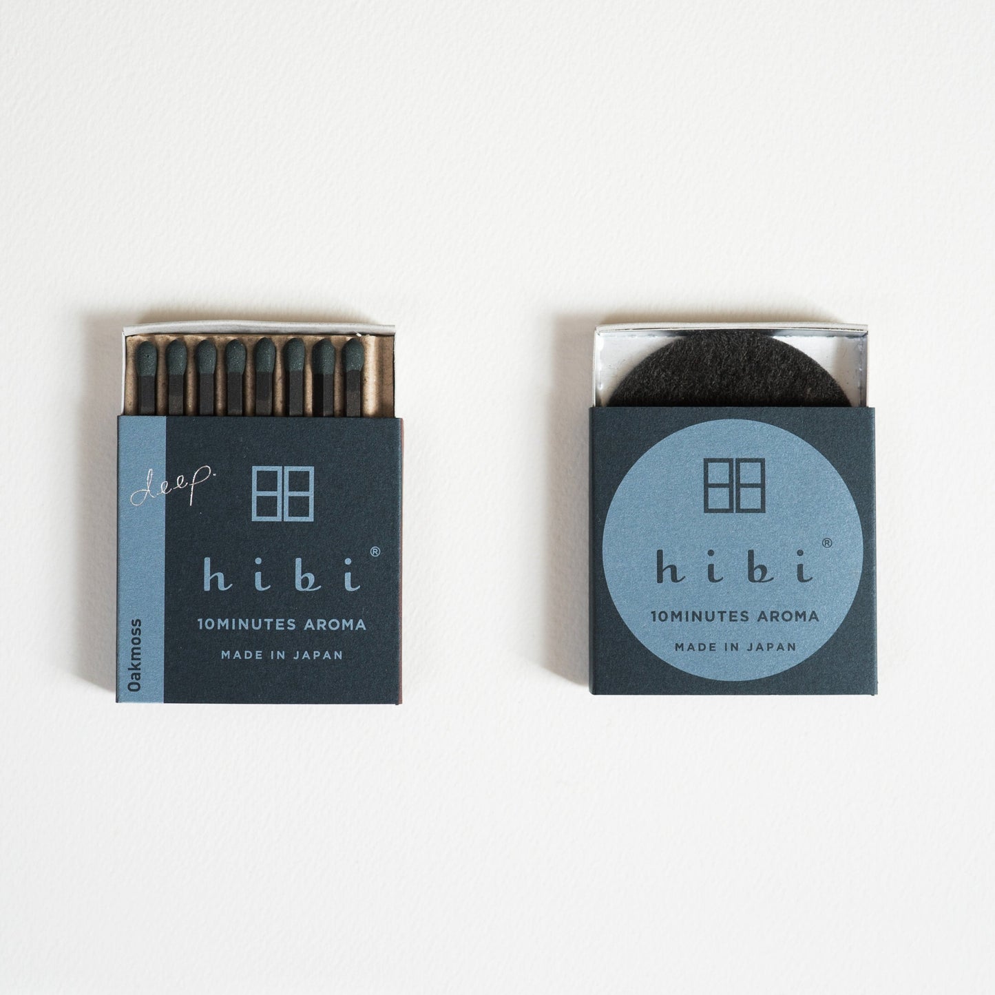 An opened box of Hibi 10 Minutes Aroma Oak Moss incense matches containing 8 incense sticks with dark green tips. Next to it is a box containing a non-flammable incense mat. All set in a white background.