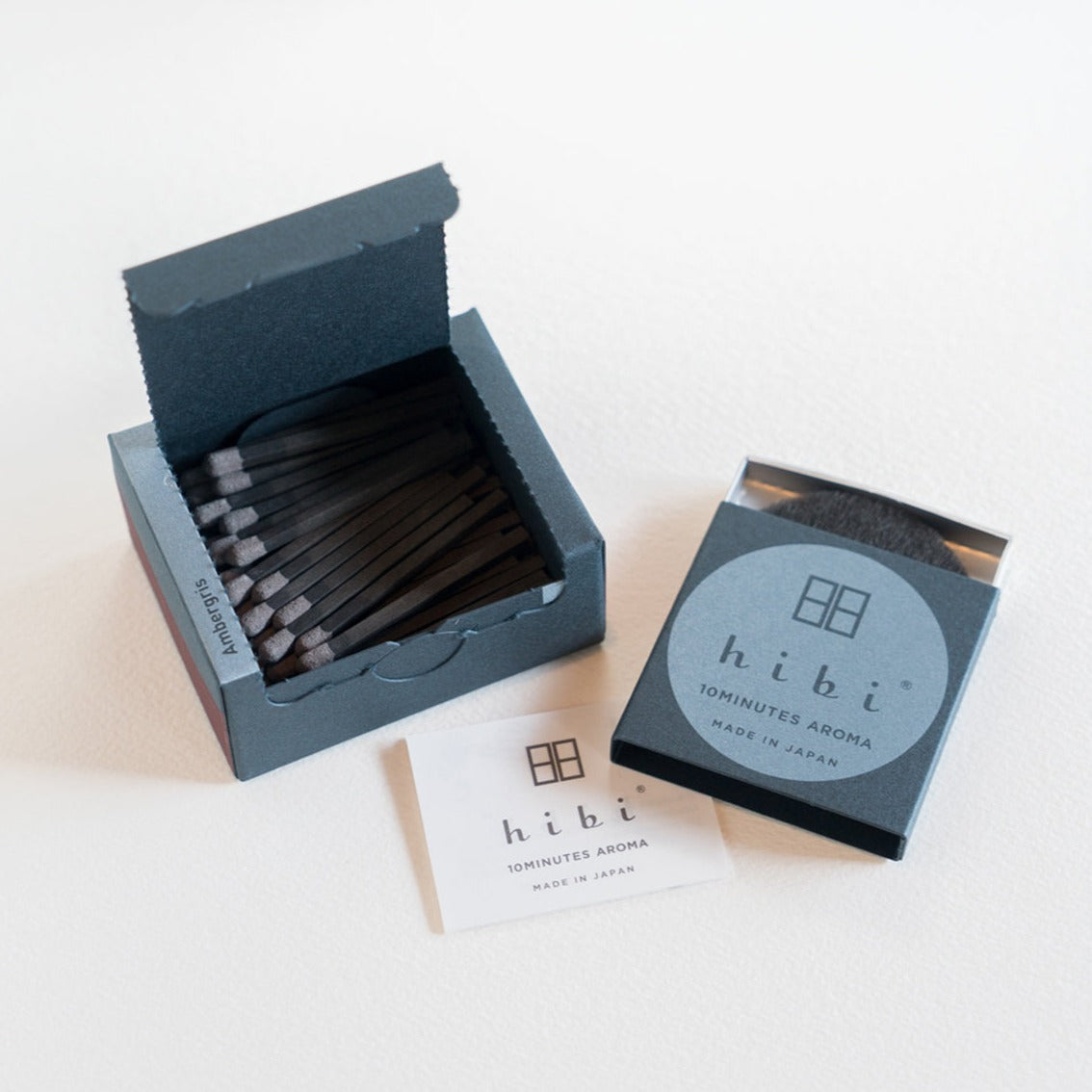 An opened box of Hibi 10 Minutes Aroma Ambergris incense matches containing 30 incense sticks with pink tips. Beside it is a box containing a non-flammable incense mat. All set in a white background.