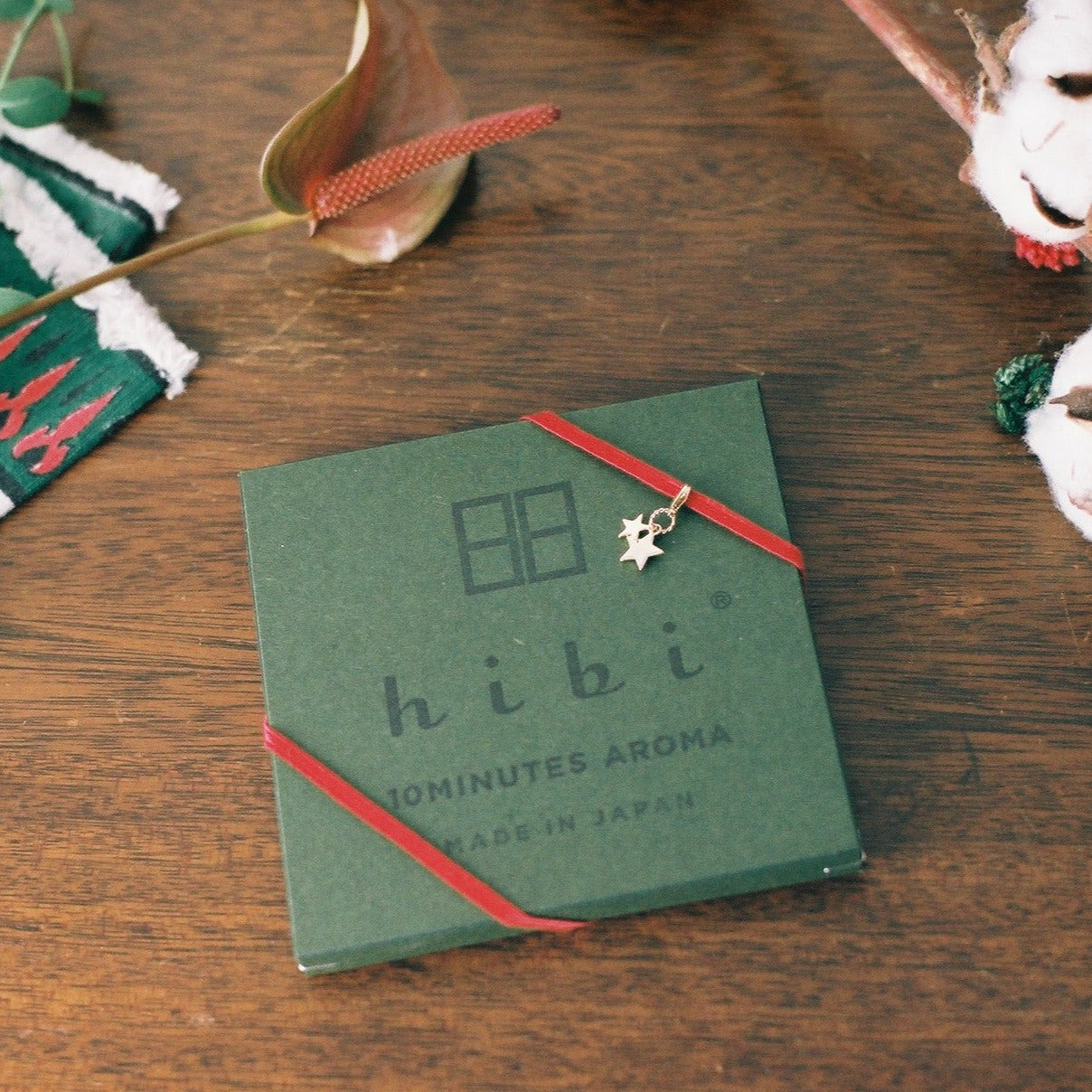 A green Hibi 10 Minutes Aroma gift box wrapped with a red ribbon and a golden star charm sits on a wood surface. Surrounding the boxes are some flowers and decorative arrangements.