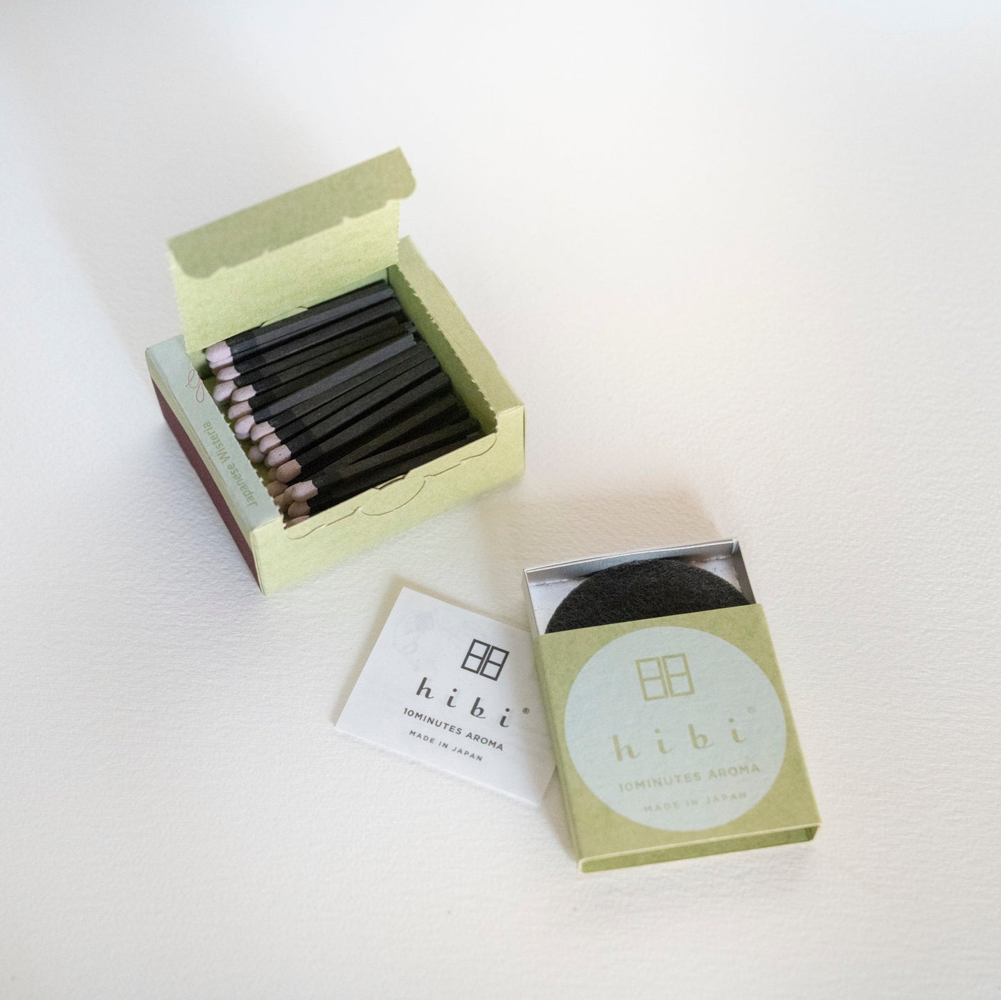An opened box of Hibi 10 Minutes Aroma Japanese Wisteria incense matches containing 30 incense sticks with pink tips, and a box containing a non-flammable mat. All set in a white background.