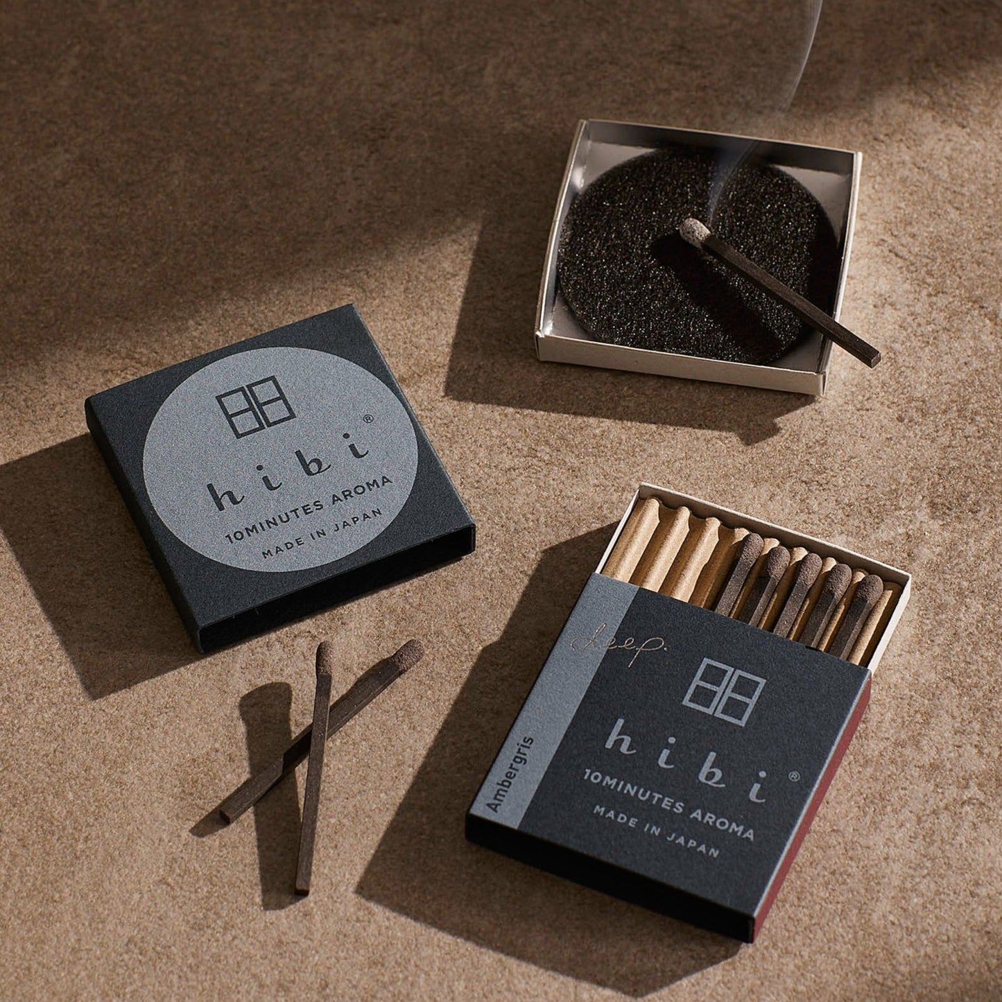 A burning incense stick resting on a non-flammable mat. Next to it is a opened box of Hibi 10 Minutes Aroma Ambergris incense matches.