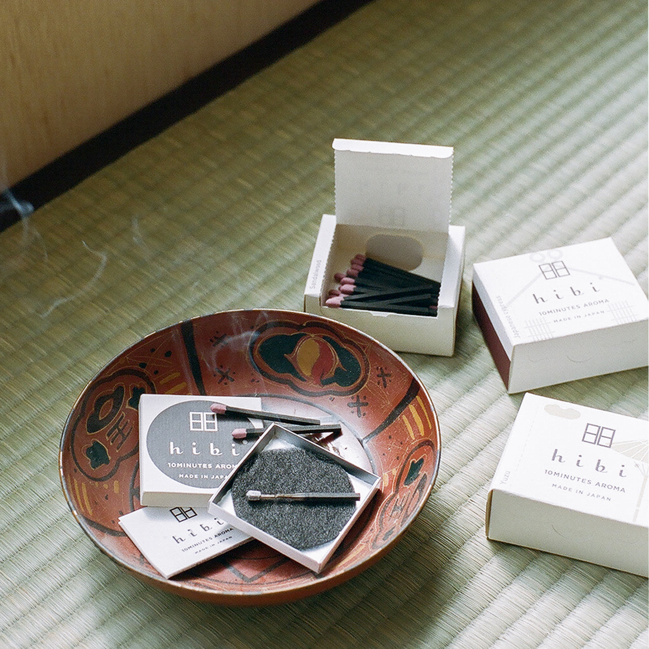 A burning incense stick is placed on top of a non-flammable mat inside a brown colour bowl, on top of a tatami mat. Three boxes of Hibi 10 Minutes Aroma incense stick, one of them opened to show 30 sticks of incense matches inside, sit next to the brown bowl.
