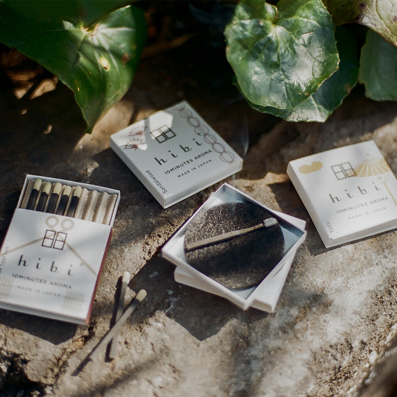 Lying on the soil are three boxes of Hibi 10 Minutes Aroma incense matches. One of them is opened and a incense stick is being burnt on a non-flammable mat.