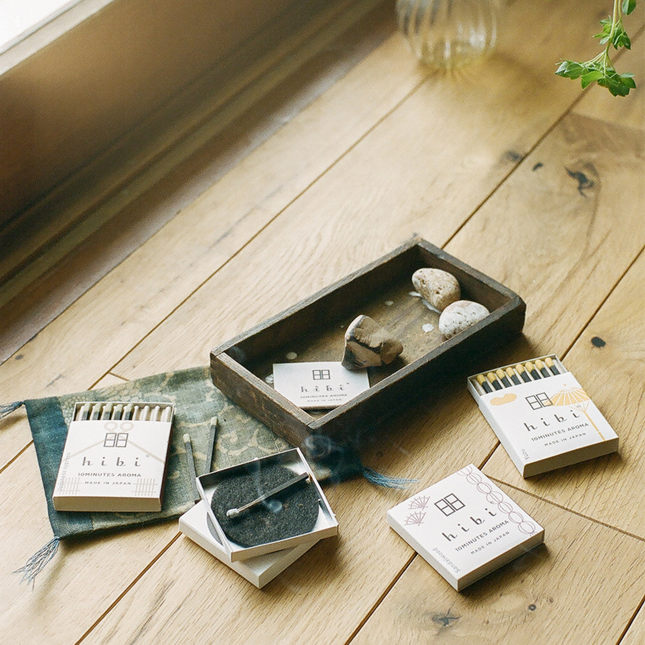 Lying on the a wood floor are three boxes of Hibi 10 Minutes Aroma incense matches. One of them is opened and a incense stick is being burnt on a non-flammable mat.