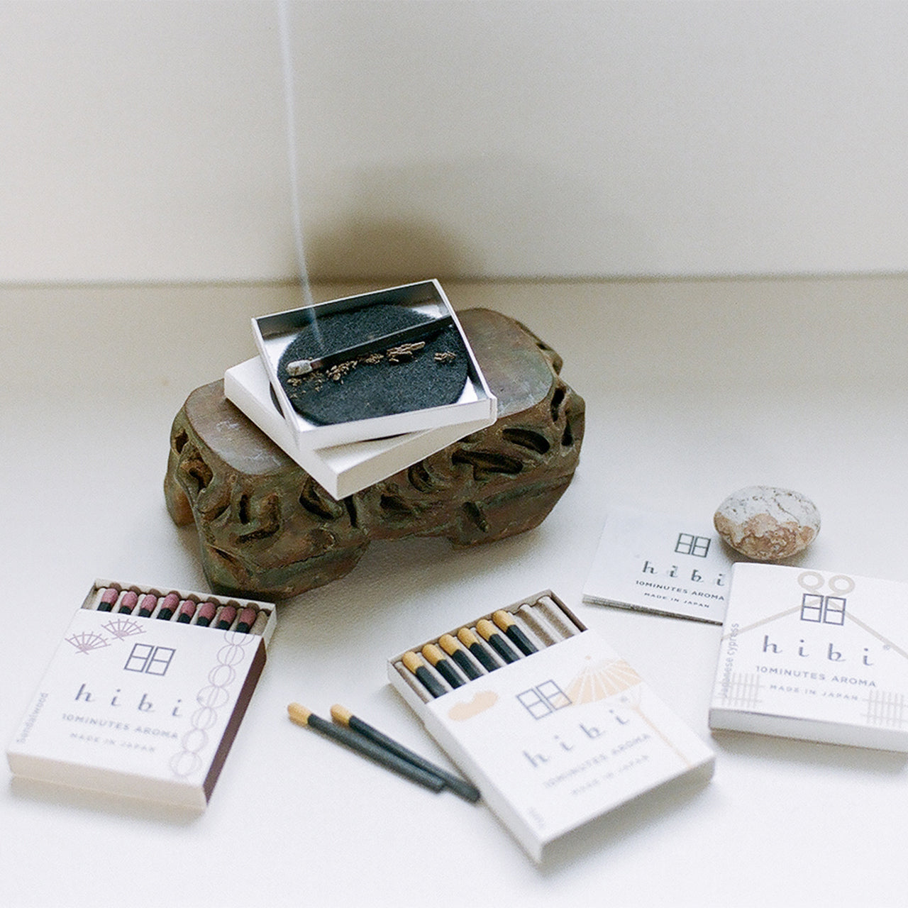 Lying on the table are three boxes of Hibi 10 Minutes Aroma incense matches. One of them are opened which show the colourful incense sticks inside. A burning incense is placed on top of a non-flammable mat over a wooden stand in the background.