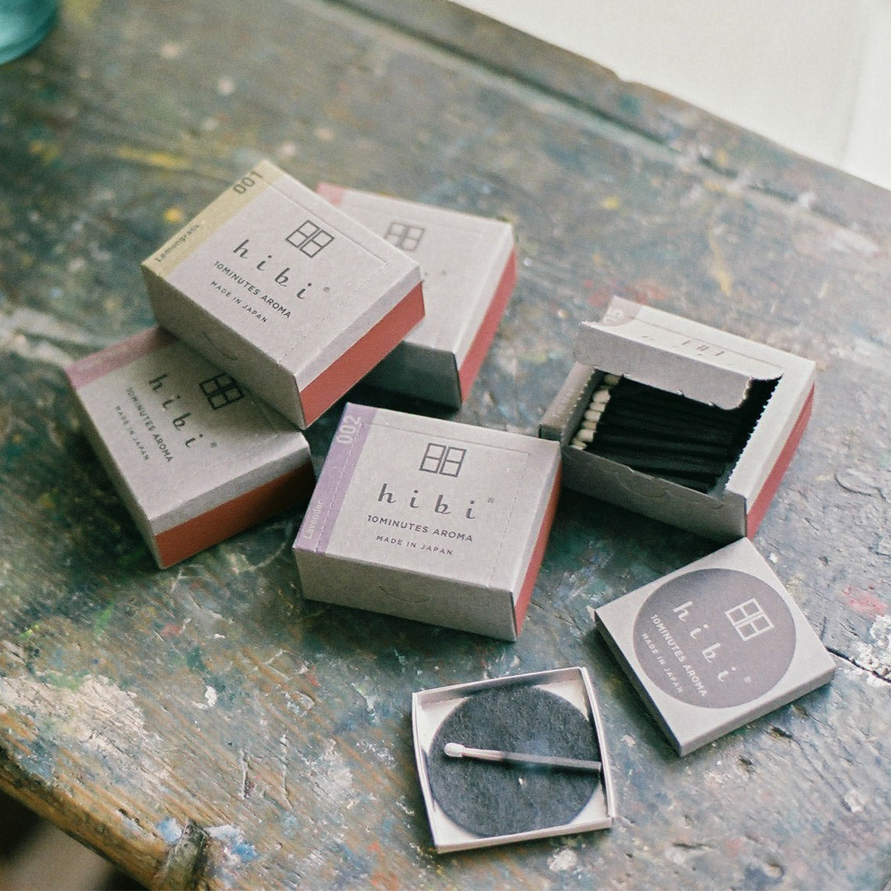 Lying on top of a wood surface are four boxes of Hibi 10 Minutes Aroma incense matches, one of them is opened and shows the white-tipped incense matches inside. Next to the incense boxes is a burning incense placed on top of a non-flammable mat.
