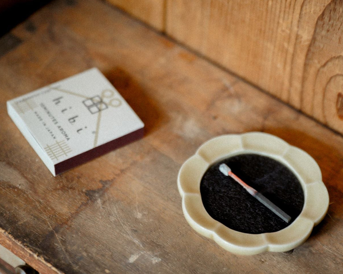 A burning incense stick on a beige flower-shape Seto potcelain tray. Next to it is a regular box of Hibi 10 Minutes Aroma incense box containing 8 incense sticks.