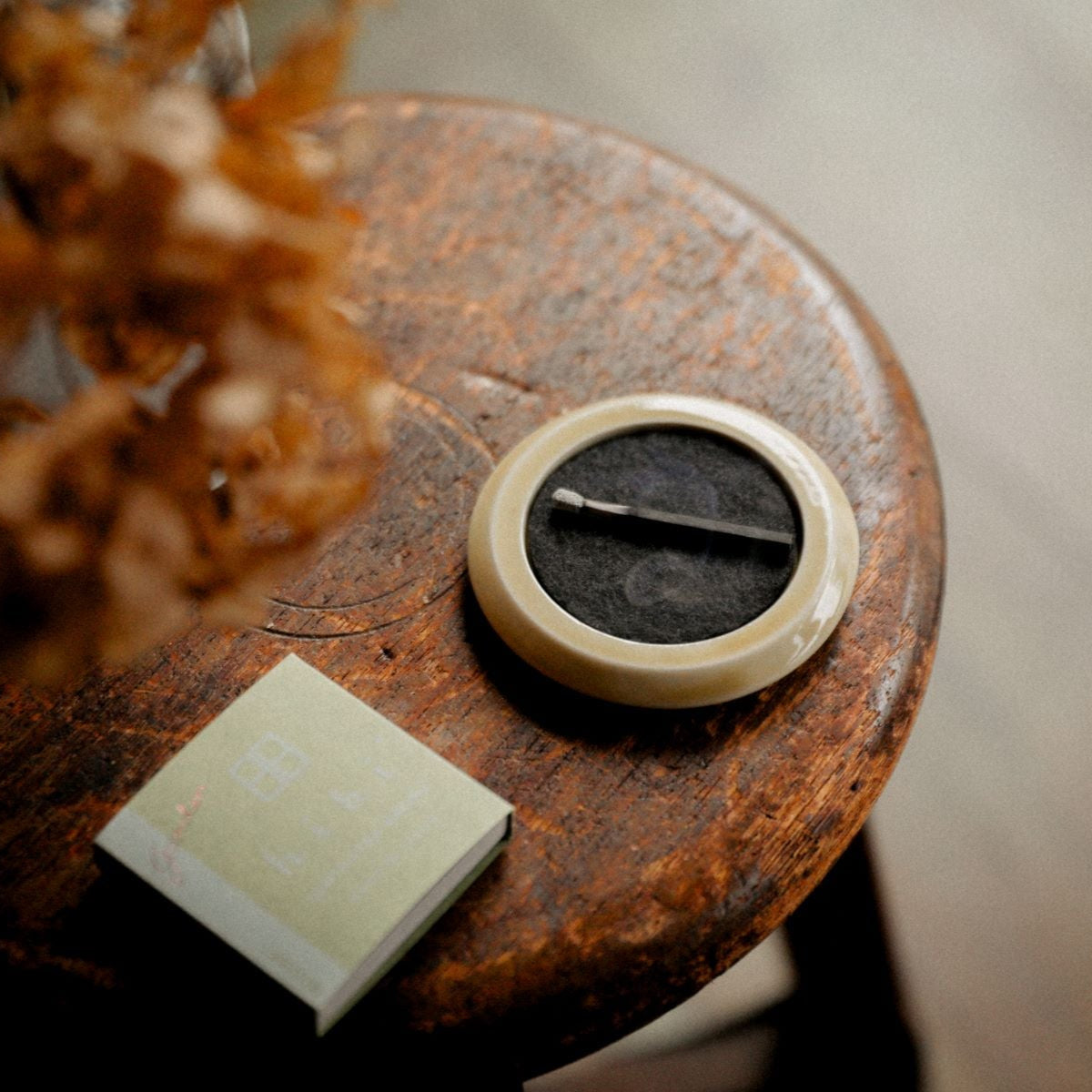 A burning incense stick on a beige round-shape Seto porcelain tray. Next to it is a regular box of Hibi 10 Minutes Aroma incense box containing 8 incense sticks.