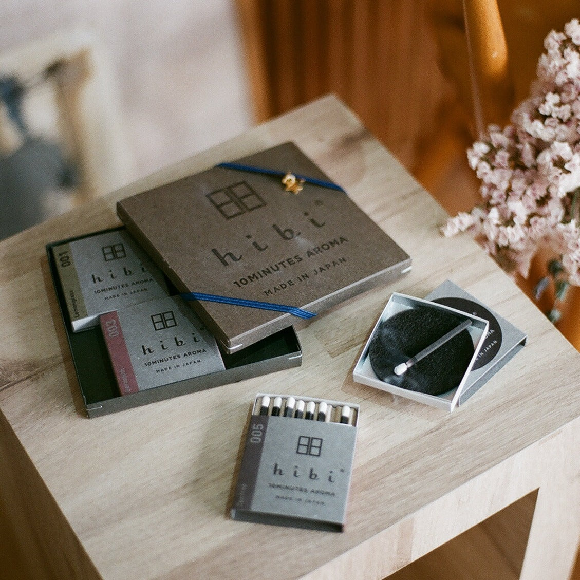 On top of a small square wooden table lies a half opened grey-colour Hibi 10 Minutes Aroma gift box. The cover of which is wrapped with a blue ribbon and a golden star charm. Inside the gift box are two small boxes of Lemongrass and Geranium incense matches. Lying next to the gift box is a small Tea Tree incense box and a burning incense placed on top of a non-flammable mat.