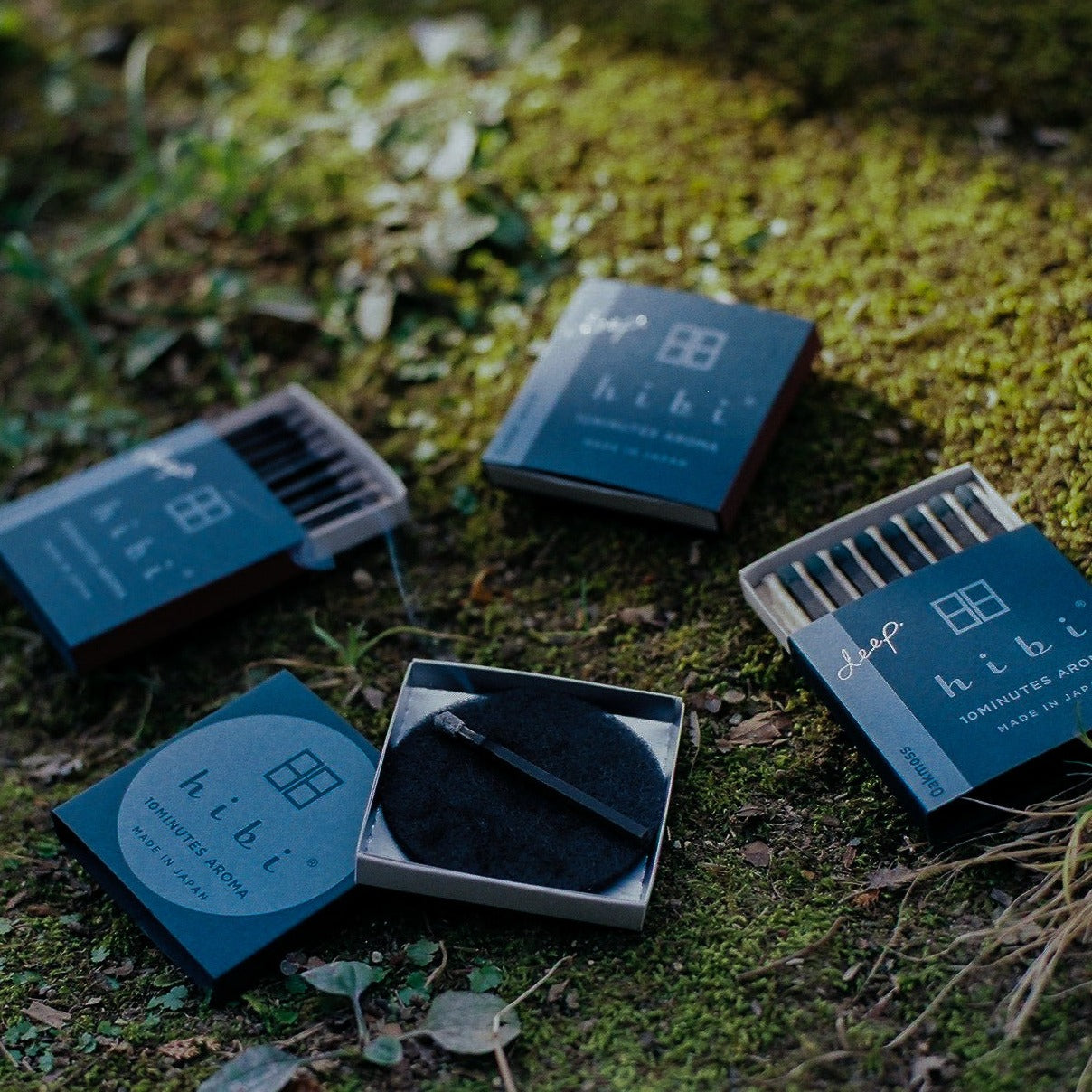 Laying on a green moss are three boxes of Hibi 10 Minutes Aroma Deep Incense matches, and a burning incense placed on top of a non-flammable mat.