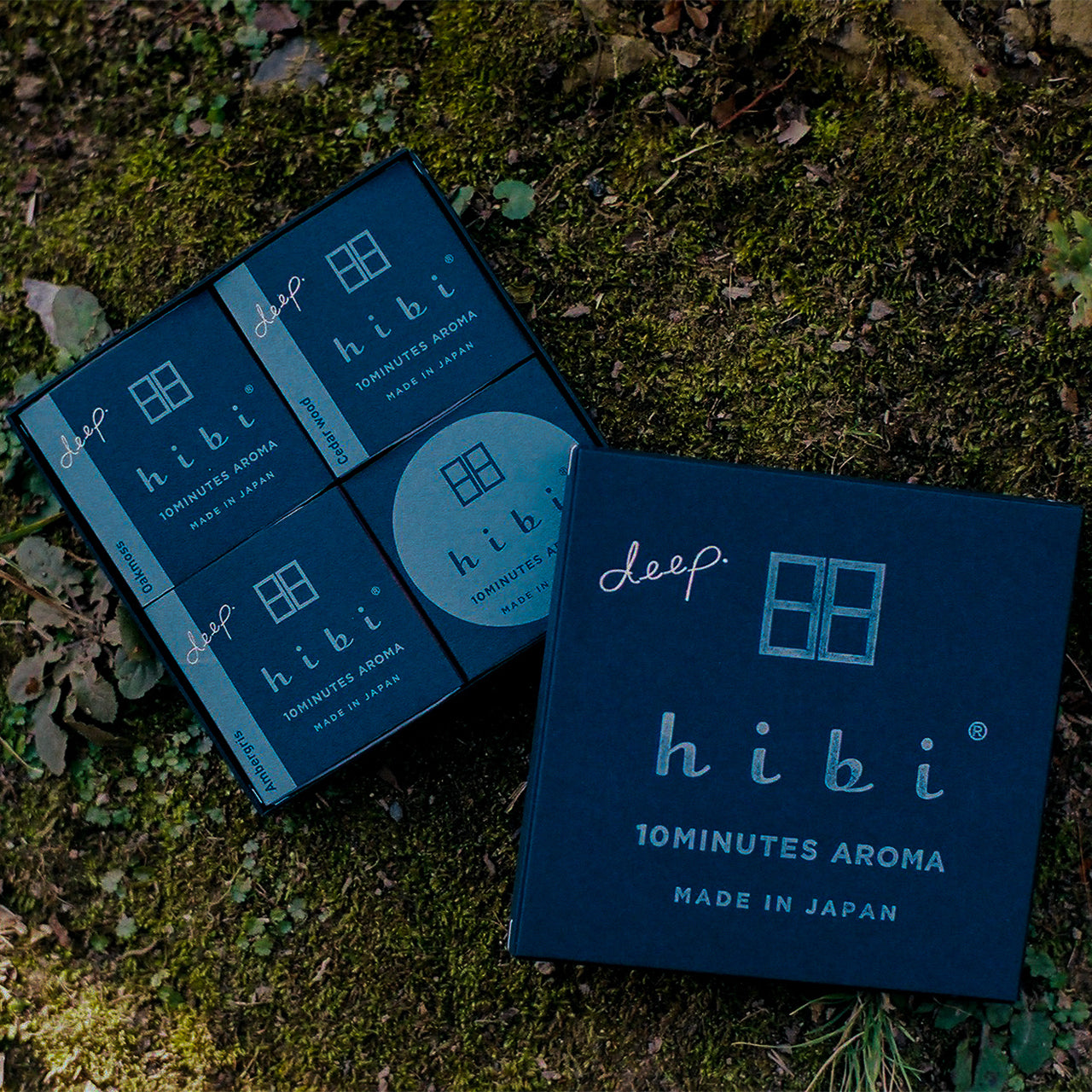 Laying on the moss ground is opened Hibi 10 Minutes Aroma gift box containing three packs of Cedar Wood, Oak Moss, and Ambergris incense matches, and a non-flammable incense mat.