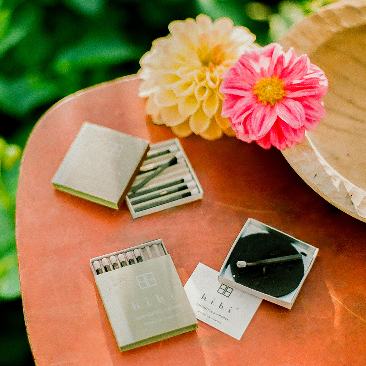 Sitting on a patio table are two opened boxes of Hibi 10 Minutes Aroma 8-stick incense matches, and a burning incense on a non-flammable mat.