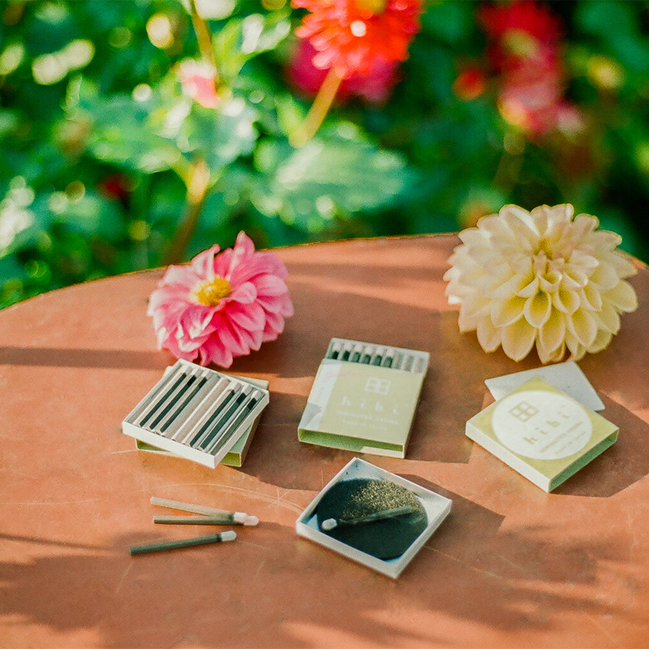 Sitting on a patio table are two opened boxes of Hibi 10 Minutes Aroma garden incense matches, a burning incense resting on a non-flammable mat, and two flowers.