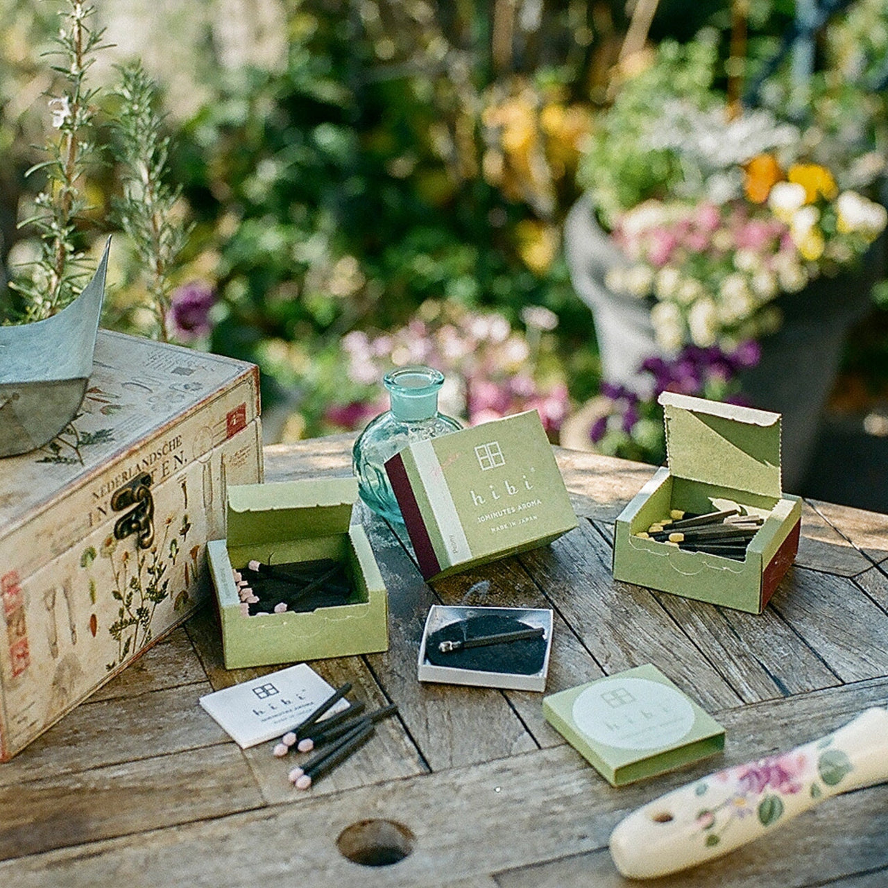 Resting on a patio table in the garden are three boxes of Hibi 10 Minutes Aroma incense matches, and a burning incense placed on a non-flammable mat . Next to them are some gardening tools.