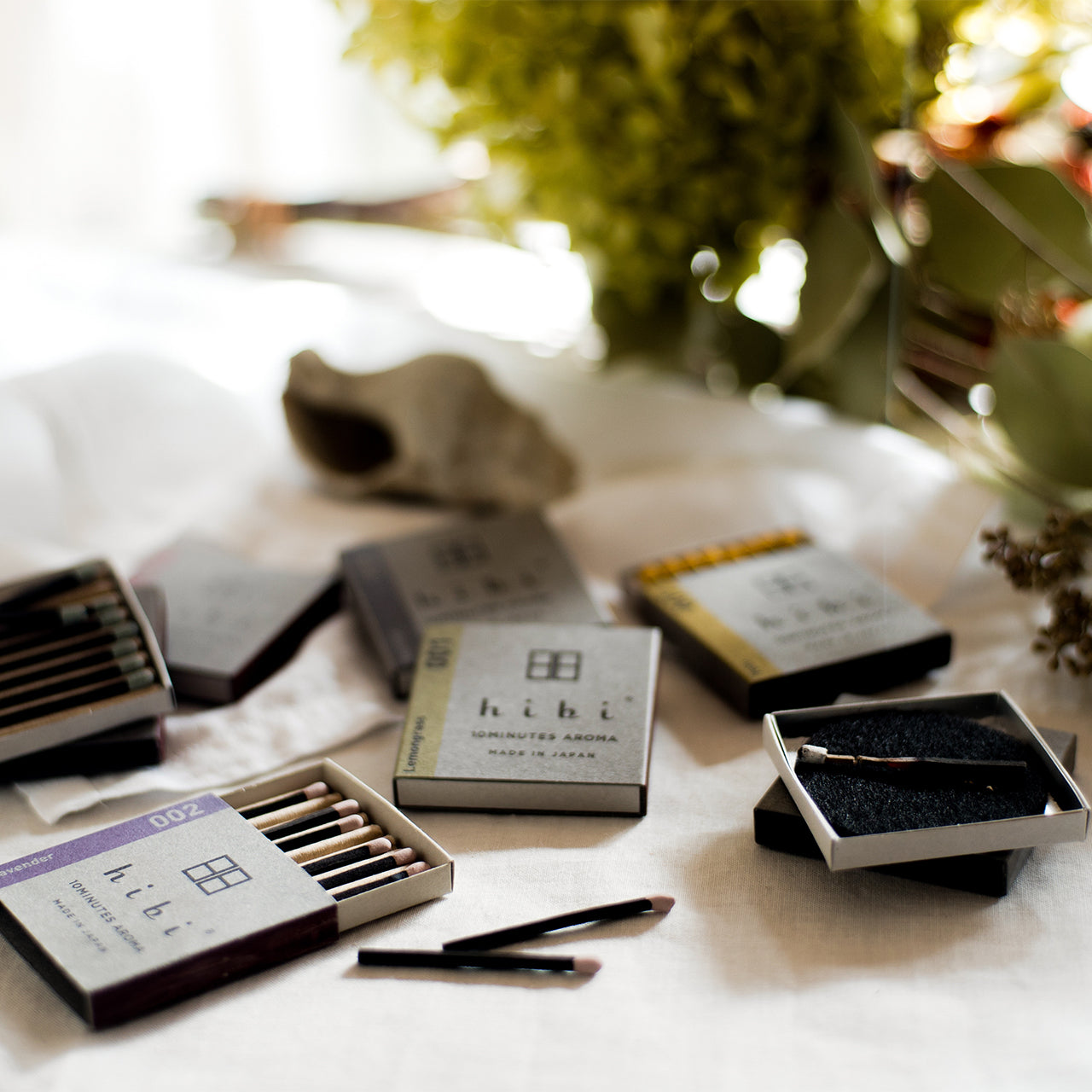 Lying on top of a table are six boxes of Hibi 10 Minutes Aroma incense matches. Two of them are opened and show the incense matches inside. Sitting next to the incense boxes is a burning incense placed on top of a non-flammable mat.