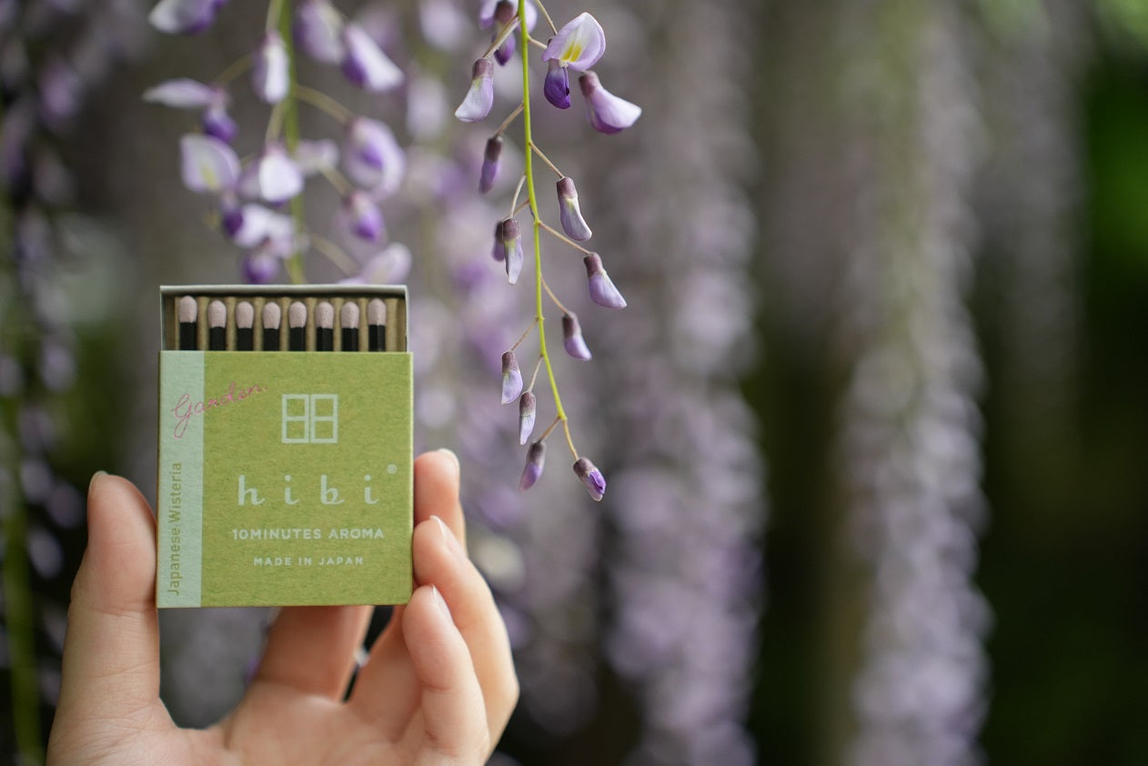 Hand holding a green hibi Japanese Wisteria incense box with purple wisteria flowers in the background