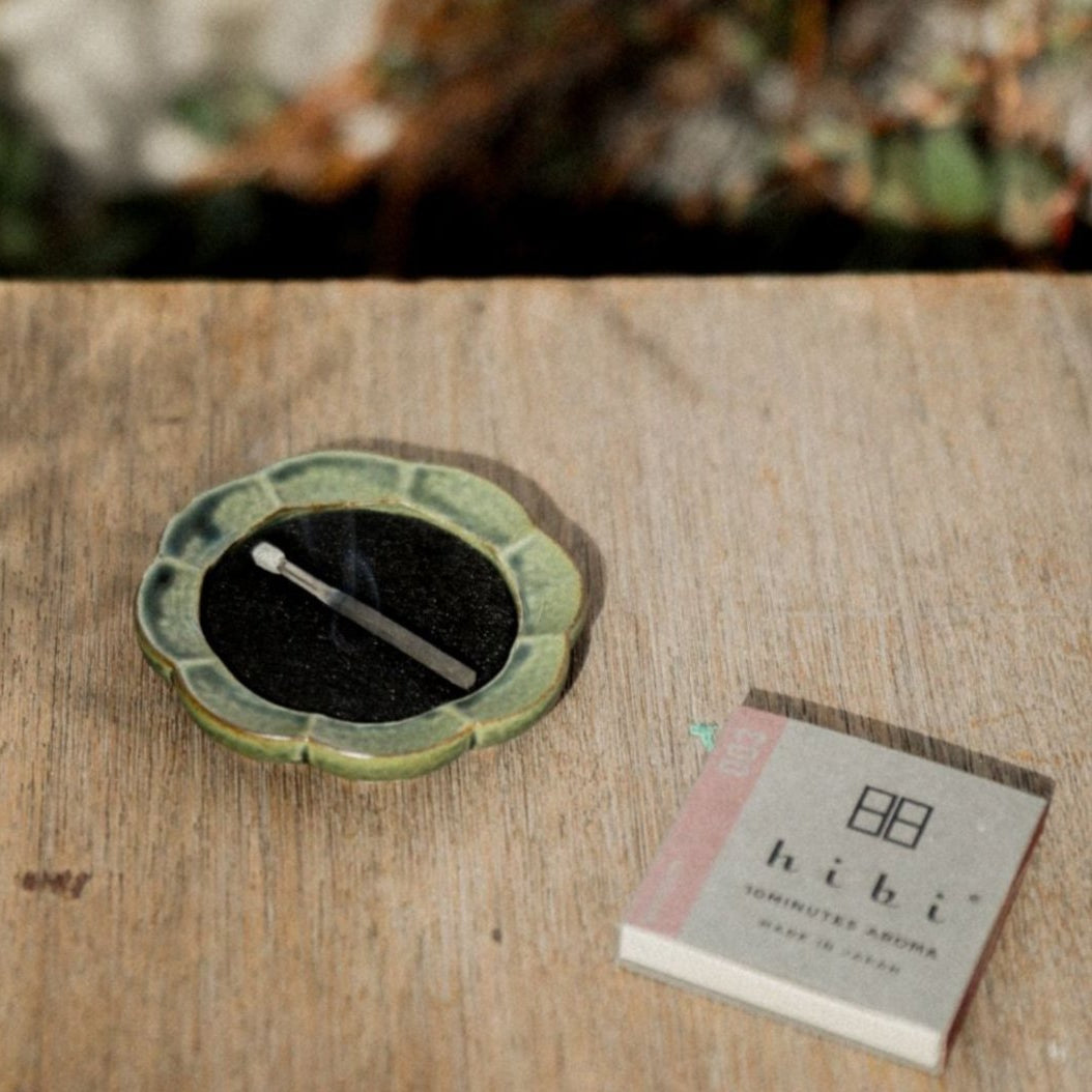 A burning incense stick on a green flower-shape Seto porcelain tray. Next to it is a regular box of Hibi 10 Minutes Aroma incense box containing 8 incense. Next to it is a regular box of Hibi 10 Minutes Aroma incense box containing 8 incense sticks.